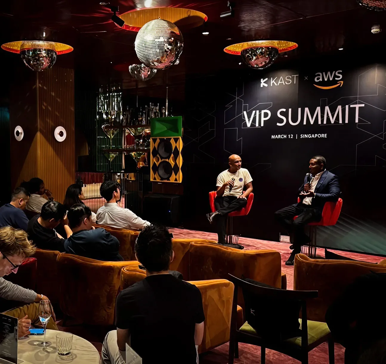 Fundadores sentados en una cumbre VIP de KAST y AWS en Singapur, viendo una conversación en el escenario.